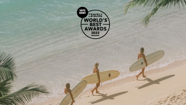 3 surfers with boards walking along beach. Travel + Leisure Worlds Best Vote Now Logo at top. 