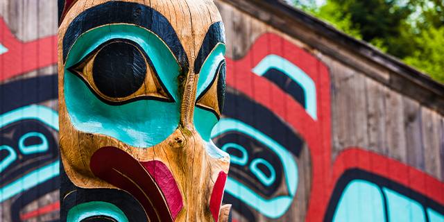 Close-up of a colorful Tlingit totem pole with traditional carving details in front of a tribal house with mural art in Alaska.